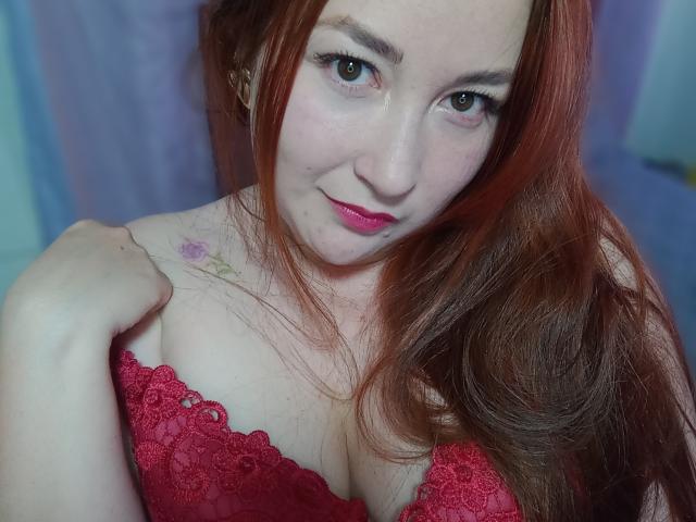 ReginaLov with red hair, pink lipstick, and a red lace bra, showing her tits with a slight smile, right hand near her shoulder, visible tattoo on upper left chest.