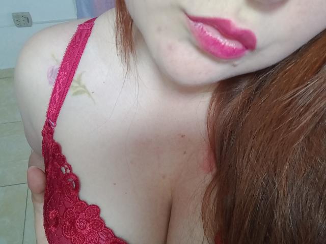 ReginaLov with red lace bra, pink lips, pale skin, and red hair, showing her tits and a small patch of cum on her shoulder in a selfie.