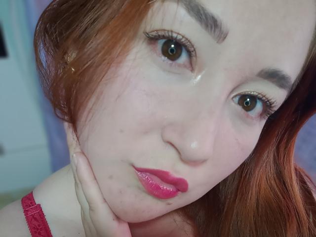 ReginaLov has pale skin, red hair, brown eyes, and pink lipstick with glossy lips, resting her hand on her chin, showing subtle freckles and a slight sheen on her face.