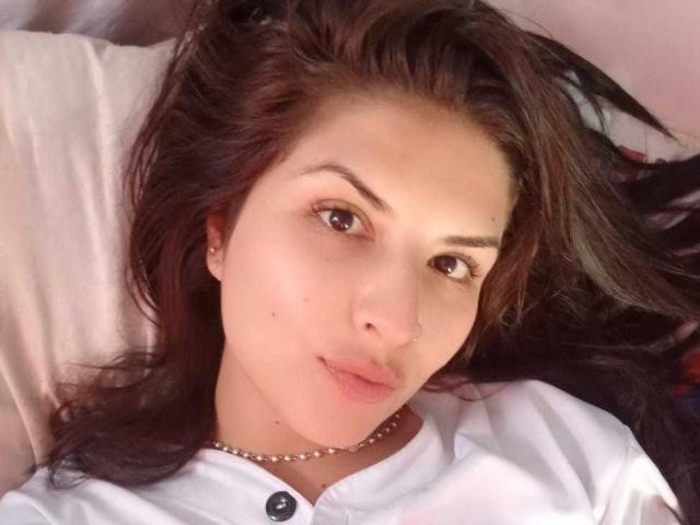 Ailinn's face, lying down, with brown eyes, dark brown wavy hair, white shirt, buttoned, small nose piercing, subtle smile, pearl necklace, light skin, and a slight pinkish hue.