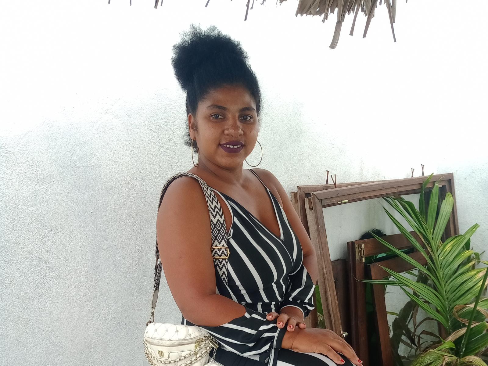 SarahRah, com pele escura, um grande cabelo afro preto, vestindo um vestido listrado preto e branco, brincos de argola e uma bolsa estampada, sentada com os seios e nádegas ligeiramente visíveis..