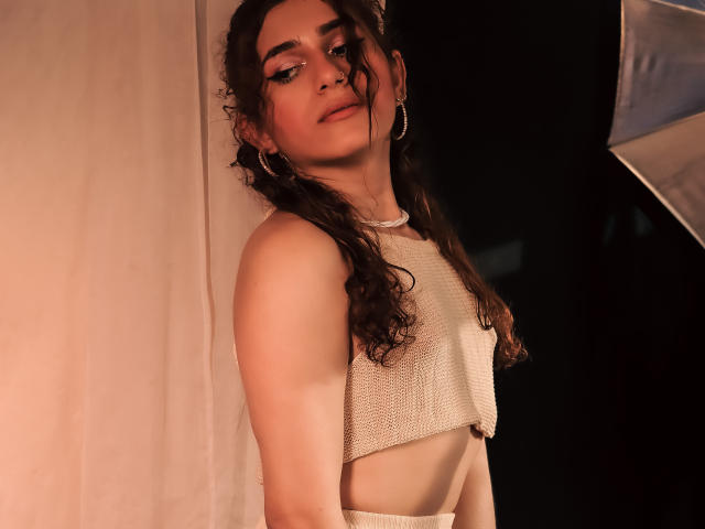 AndyDupont, avec ses cheveux bruns bouclés, portant un haut en mesh blanc, mettant en valeur une silhouette fine et de petites seins, avec des boucles d'oreilles cerceaux et un sourire discret, dans une lumière douce..