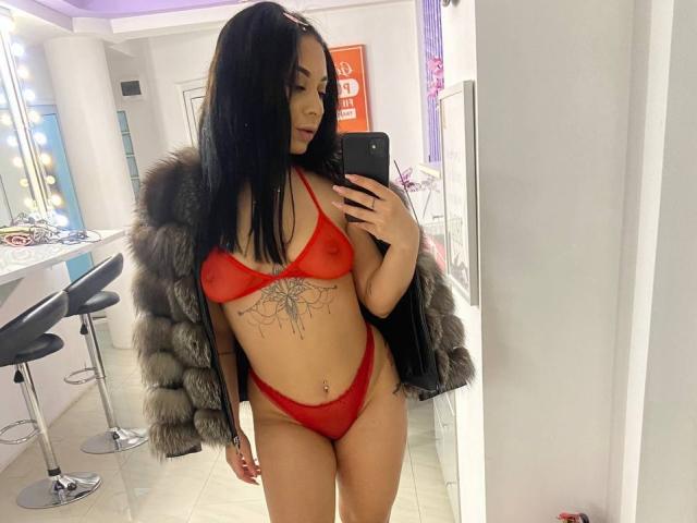 MadyLux mit schwarzen Haaren, trägt einen roten, durchsichtigen BH und einen String, eine graue Felljacke, zeigt ein Tattoo auf ihrem Bauch, hält ein Telefon, steht vor einem Spiegel und macht ein Selfie, wobei ihre kleinen Brüste und ihr Hintern zu sehen sind..