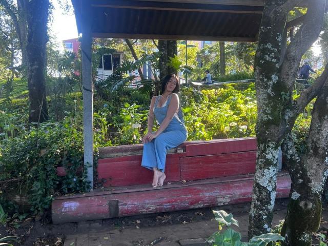 NicoColeman, with curly brown hair, sits on a red wooden bench, wearing a light blue dress, barefoot, under a wooden shelter, surrounded by greenery and trees.