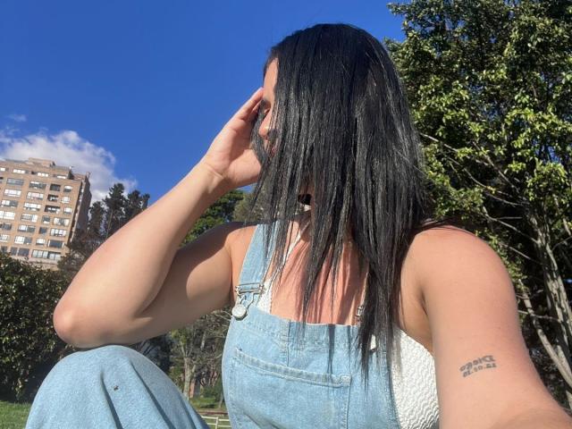 NicoColeman with black straight hair, wearing denim overalls and white tank top, touching face with right hand, visible tattoo on left arm, blue sky background.