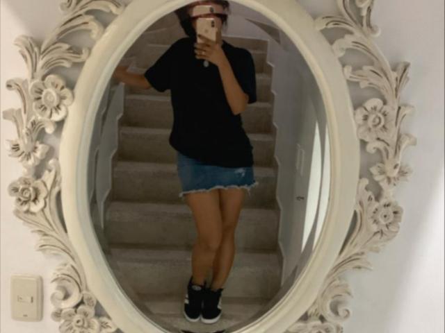 CaterinaRomano with tan skin, black t-shirt, blue denim shorts, and black sneakers, standing in front of an ornate mirror, holding a phone, showing her ass and legs.