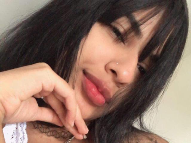 PamelaHotCouple's face, slightly angled, with black hair, bangs, and a small nose piercing. She's smiling with pink lips, hand near chin, and a tattoo visible on her neck.