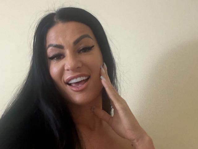 AndraD with long black hair, thick eyebrows, and long eyelashes, smiling with tongue out, touching her cheek with a hand, showing off her white teeth and pink lips.