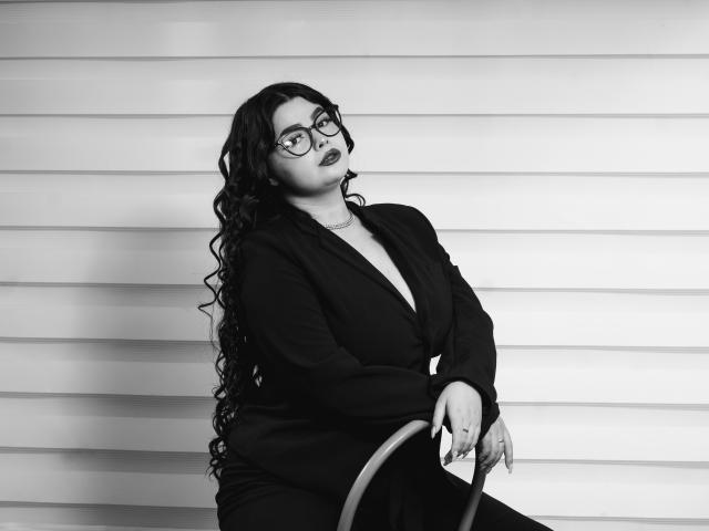 Foto in bianco e nero di BrithneyLee con lunghi capelli neri ricci, occhiali e rossetto scuro, che indossa un blazer nero, seduta, con le mani appoggiate sulla sedia, che guarda verso l'alto, mostrando leggermente il sedere..