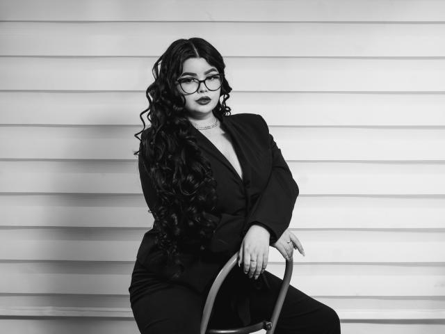 Black and white photo of BrithneyLee with long curly hair, glasses, dark lipstick, black blazer, and pants, sitting on a chair, hand on armrest, looking at camera.