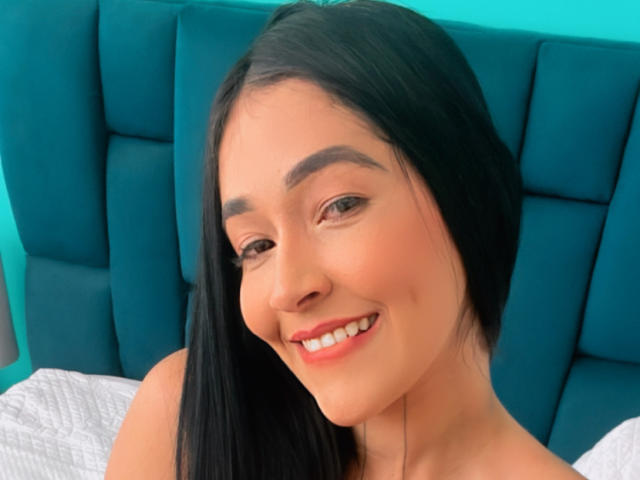 NicoColeman has long black hair, brown eyes, and is smiling with red lipstick. She's topless, showing a cute face and small tits, against a teal headboard.