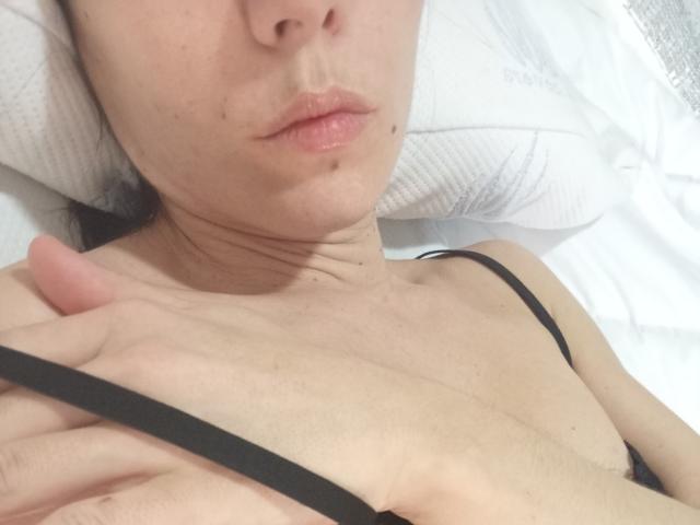 OrianaLaFrancaise liegt auf einem weißen Kissen, mit leicht geschürzten Lippen, heller Haut, einem schwarzen BH-Bügel, einer Hand auf der Brust, sichtbaren Halsfalten und kleinen Sommersprossen auf der Wange, mit dezentem Make-up..