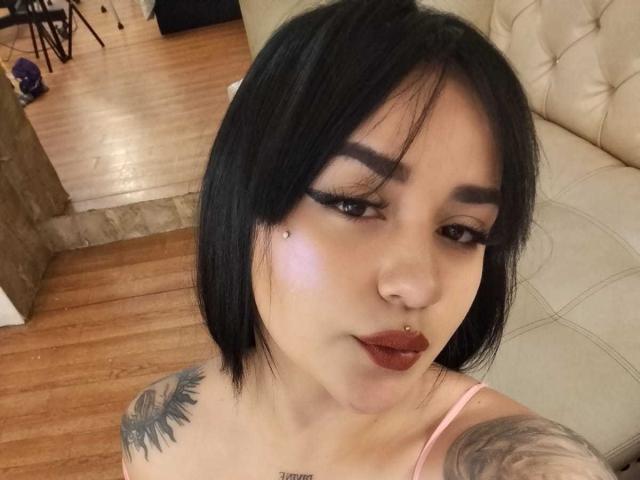 KarinaBlake has black hair, thick eyebrows, eyeliner, and a nose piercing. She's wearing dark lipstick and has tattoos on her shoulder. Her pink top shows her tits.