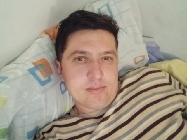 MysteriousMan69 tem cabelo preto curto, olhos castanhos e uma expressão neutra, deitado na cama com um travesseiro e cobertor listrados, seu rosto está quase todo visível, ligeiramente coberto por um cobertor com listras marrons..