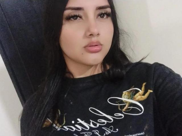 AmaliaDalton has black hair, brown eyes, and pink lips with a small mole on her right cheek. She wears a black shirt with gold embroidery.