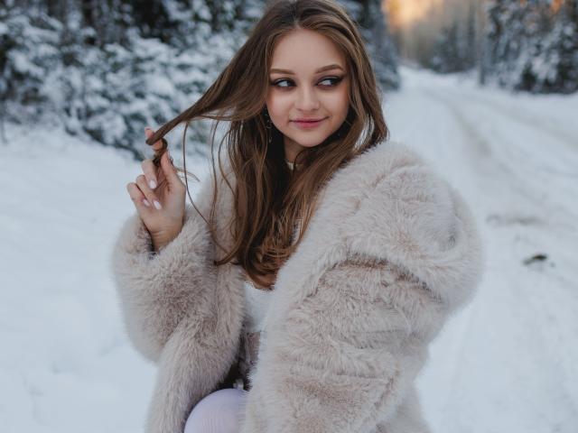 MayxFlower with long brown hair, wearing a fluffy beige coat, holding her hair, smiling, snow-covered forest path in background, pink lips, white top, looking to the side.