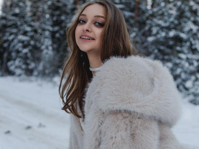 MayxFlower with long brown hair, wearing a fluffy beige fur coat, smiling in a snowy forest. Her pink lips and subtle makeup are visible. Her tits are partially covered.