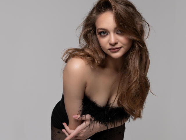 MayxFlower with long brown hair, black feathered top, and black lace stockings, leaning forward, showing her tits, with a subtle smile, and soft makeup, against a gray background.