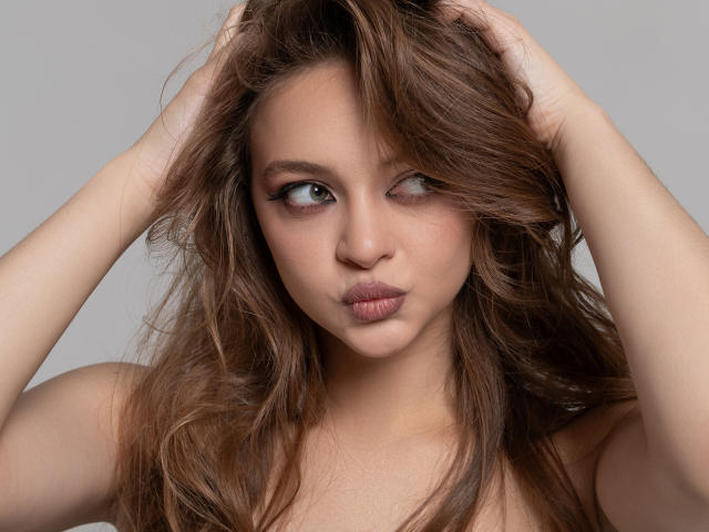 A MayxFlower tem cabelos castanhos ondulados, lábios rosados e está olhando para o lado, com as mãos no cabelo, exibindo seus seios e seus olhos castanhos sensuais..