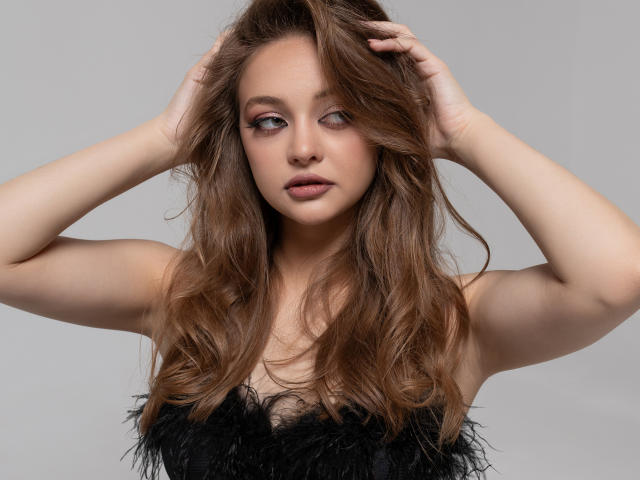 MayxFlower has long, wavy brown hair, fair skin, and wears a black feathered top, with hands in her hair, looking slightly to the side with a neutral expression.