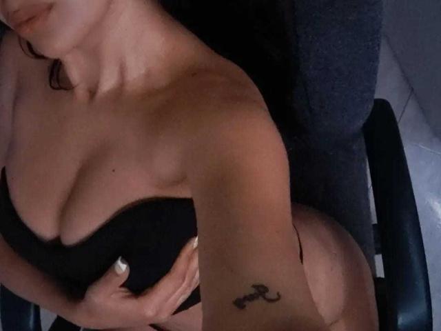 AndreaPretty with tan skin, black hair, holding black bra covering her tits, red thong, tattoo on right arm, sitting on dark chair, visible collarbone, white nail polish.