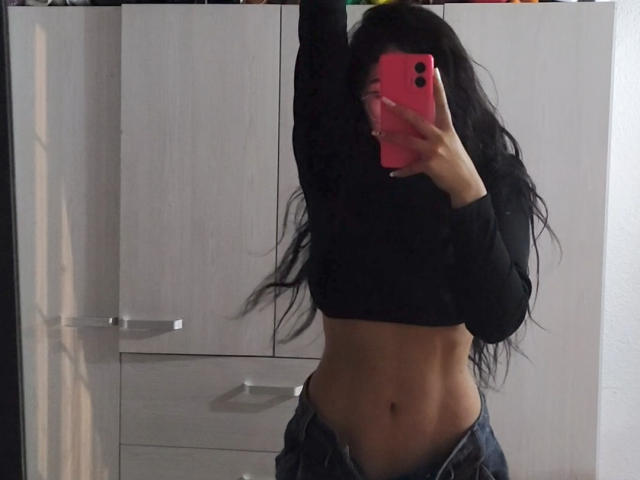 Andrea, avec de longs cheveux noirs, portant un crop top noir, dévoilant ses abdominaux et un jean déboutonné, prend un selfie devant un miroir avec un téléphone rose..