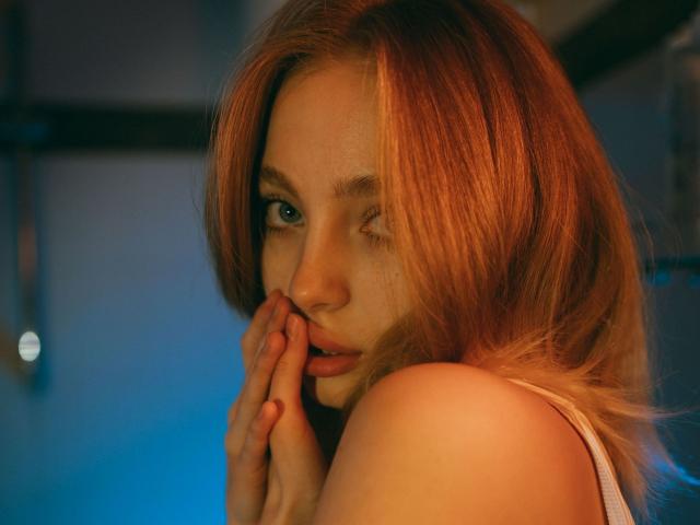 LisaMetalHoney con i capelli rossi ondulati, un occhio chiuso, una mano vicino alla bocca, una luce blu e arancione soffusa, indossa un canottino bianco, mostrando il sedere e con un'espressione delicata..