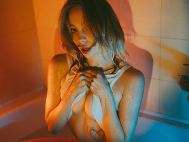 LisaMetalHoney mit feuchter, glänzender Haut, die ein weißes Hemd über ihrer Brust hält, blaues und oranges Licht, kurze blonde Haare, Tattoo an ihrer rechten Brust, blickt in die Kamera..
