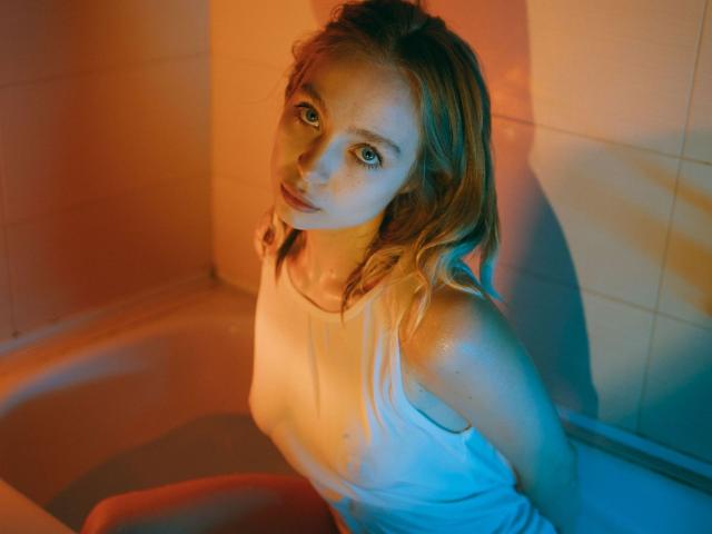 LisaMetalHoney con canottiera bianca bagnata, seduta nella vasca da bagno, illuminazione blu e arancione, che guarda verso l'alto con espressione seria, capelli biondi, piccoli seni, figura snella, lentiggini, goccioline d'acqua sulla pelle..