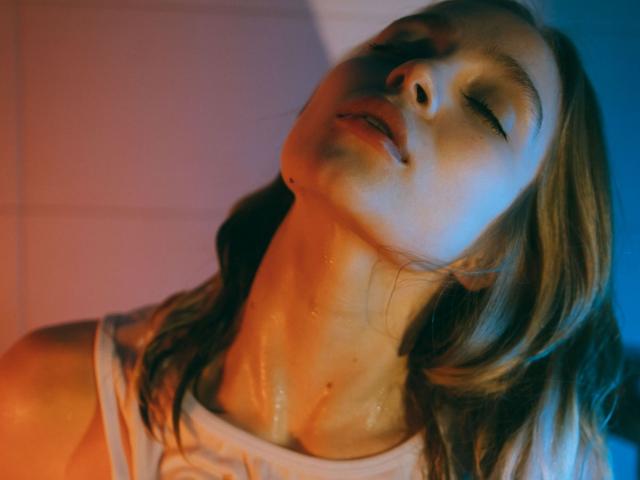 LisaMetalHoney's face is tilted back with eyes closed, wet skin glistening, wearing a white tank top. Orange and blue lighting highlights her neck and chin.
