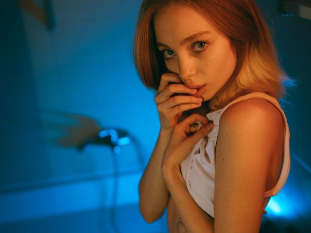 LisaMetalHoney with short, blonde hair, biting her finger, wearing a white tank top, exposing her small tits, in a blue-lit room, looking seductively at the camera.