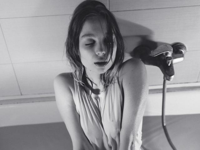 Black-and-white photo of LisaMetalHoney with wet, shoulder-length hair, eyes closed, wearing a soaked, sleeveless top, holding a showerhead, water droplets on her skin, and a relaxed expression.