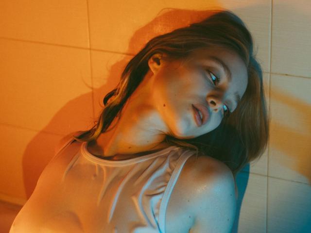 Lisa, con una canottiera bianca bagnata, appoggiata al muro, illuminata da luci arancioni e blu, con la testa inclinata, gli occhi socchiusi, i capezzoli visibili, un'ombra sulla guancia, le labbra leggermente dischiuse..