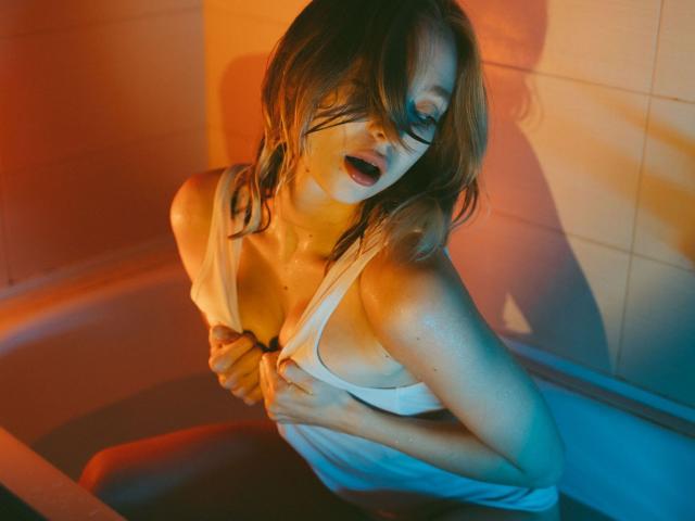 LisaMetalHoney with wet, white tank top, holding her tits, mouth open, orange and blue lighting, sitting in a tub, shadow on wall, wet skin, messy brown hair.