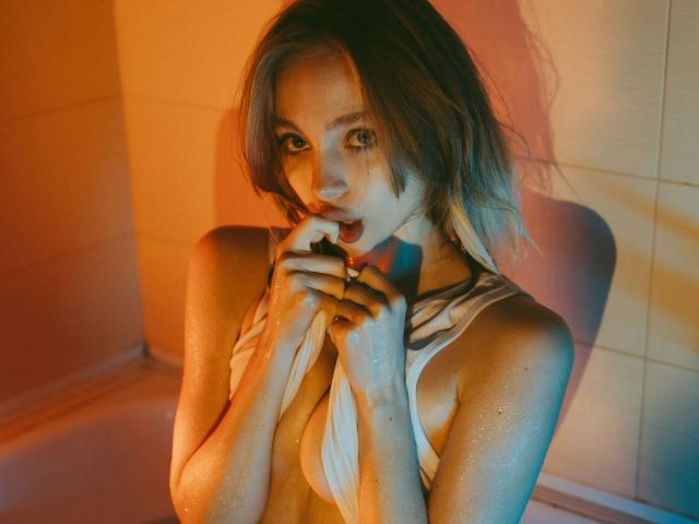 LisaMetalHoney, con la pelle umida, i capelli biondi e gli occhi verdi, che si morde un dito, indossa una maglietta bianca bagnata, che mette in risalto il suo seno piccolo e il suo corpo snello e sexy..