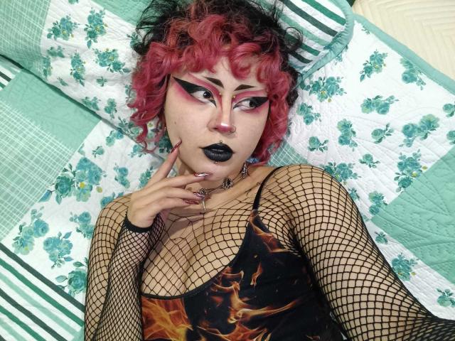 GothVioleth with curly pink hair, black eyeliner, pink eyeshadow, black lipstick, fishnet sleeves, flame-patterned top, lying on a green and white quilted bed, touching her chin.