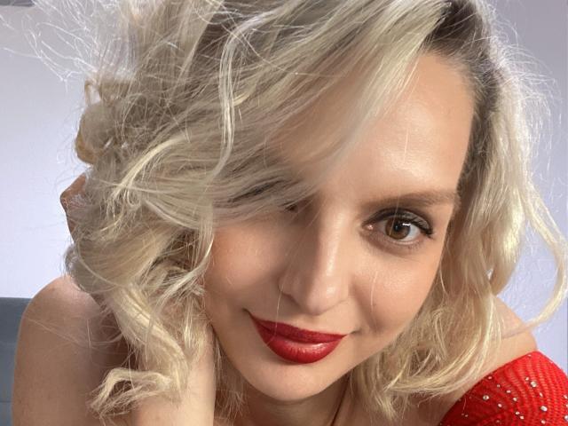 DouceAmelia has blonde curly hair, red lipstick, and a cute smile. She's wearing a red, sparkly top, looking directly at the camera with brown eyes.