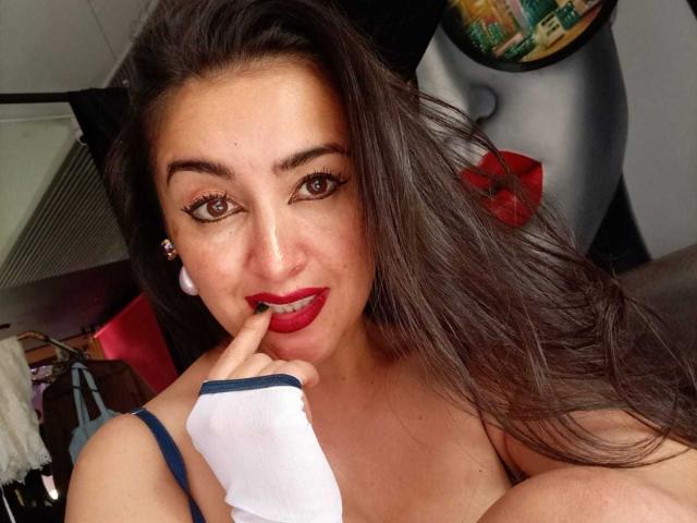 CherryHott69 with red lipstick, brown eyes, and long dark hair, touching her red-lipped lips with a finger, wearing a white glove and blue strap. Boobs visible.