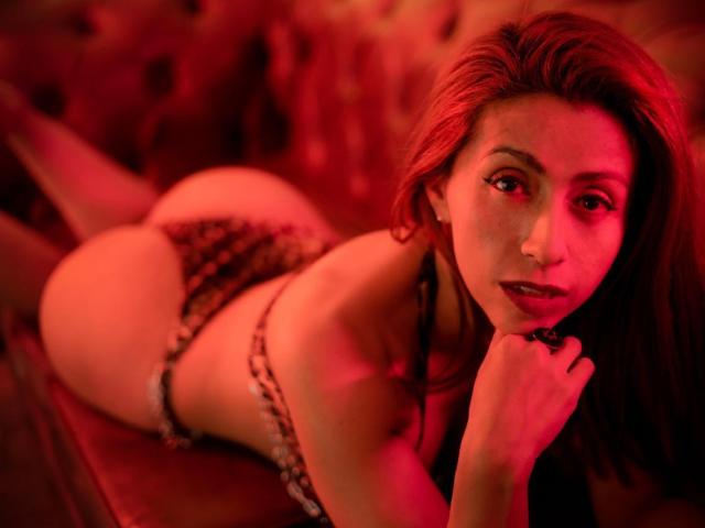 LauraBluess lying on a red couch, wearing leopard print lingerie, showing her ass, looking at the camera with red lighting. Her hand is near her mouth, she has red hair.