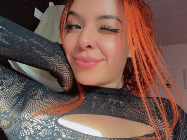 TammyyRoss, avec des cheveux orange, portant un haut en dentelle noire qui dévoile sa poitrine, clignant de longs cils, souriant à la caméra, une main derrière sa tête..