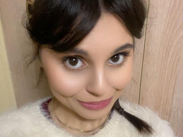 MeEleonora has medium brown eyes, dark brown hair in a braid, pink lipstick, and a cute smile, wearing a fluffy white top with a gold chain necklace.