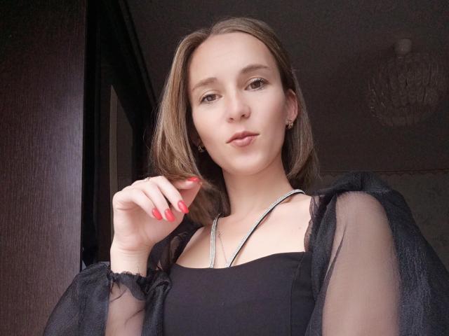 SueShine with medium brown hair, black sheer top, red nails, silver necklace, looking at camera, hand near face, subtle smile, chandelier in dark room, soft lighting.