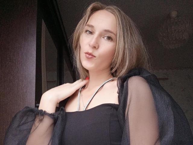 SueShine with medium tits, wearing a sheer black top, has light brown hair, red nails, and a silver necklace, looking slightly to the side with a pouty expression.