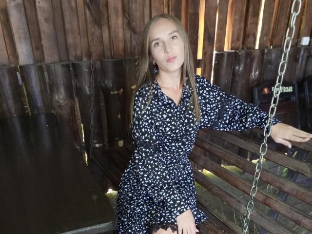 SueShine with long brown hair, wearing a blue floral dress, sits on a wooden swing, holding a chain, looking forward with a slight smile, in a wooden shed.