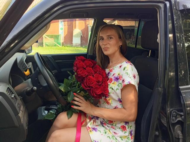 SueShine with red roses, floral dress, and pink garter, sitting in a black car, holding bouquet, looking cute, brown hair, slight smile, right hand on steering wheel.