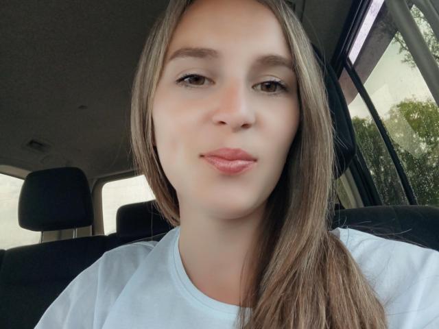 SueShine with long brown hair, light skin, and full pink lips, wearing a white shirt, sitting in a car, looking at the camera with a slight smile.