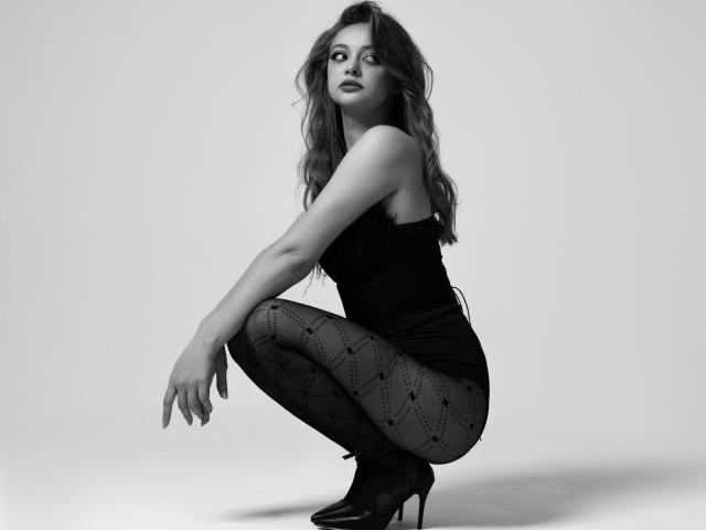 MayxFlower está agachada, vistiendo un top negro sin tirantes, medias negras estampadas y tacones altos, con cabello oscuro y ondulado, mirando hacia un lado en una foto en blanco y negro..