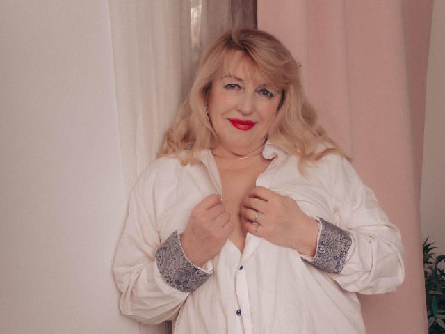 SamanthaLala mit blonden Haaren, rotem Lippenstift und einem weißen Hemd mit Knopfleiste, das einen Ausschnitt zeigt, mit den Händen auf der Brust, lächelnd, ein rosa Vorhang im Hintergrund. Brüste sichtbar, Hemd leicht geöffnet..