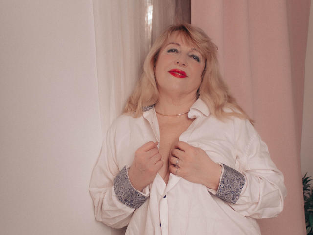 SamanthaLala with blonde hair, red lipstick, white shirt, grey cuffs, holding shirt open, showing cleavage, standing against pink curtain, smiling.