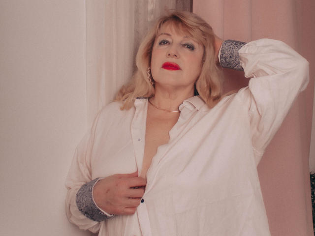 SamanthaLala has blonde hair, red lipstick, and a slightly open white shirt, revealing her small tits, with one hand in her hair and the other on her chest.